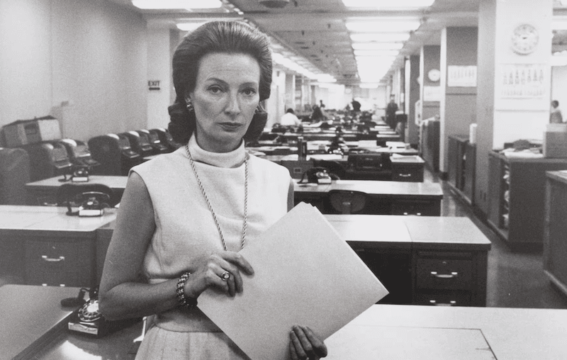 Charlotte Curtis in the New York Times newsroom. She is a smartly dressed 1960s woman. She is holding a thin folder containing a news story or something more.