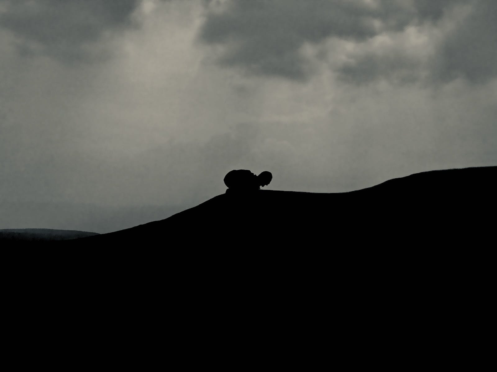 A strange figure crouches on a hillside with menacing clouds behind them.