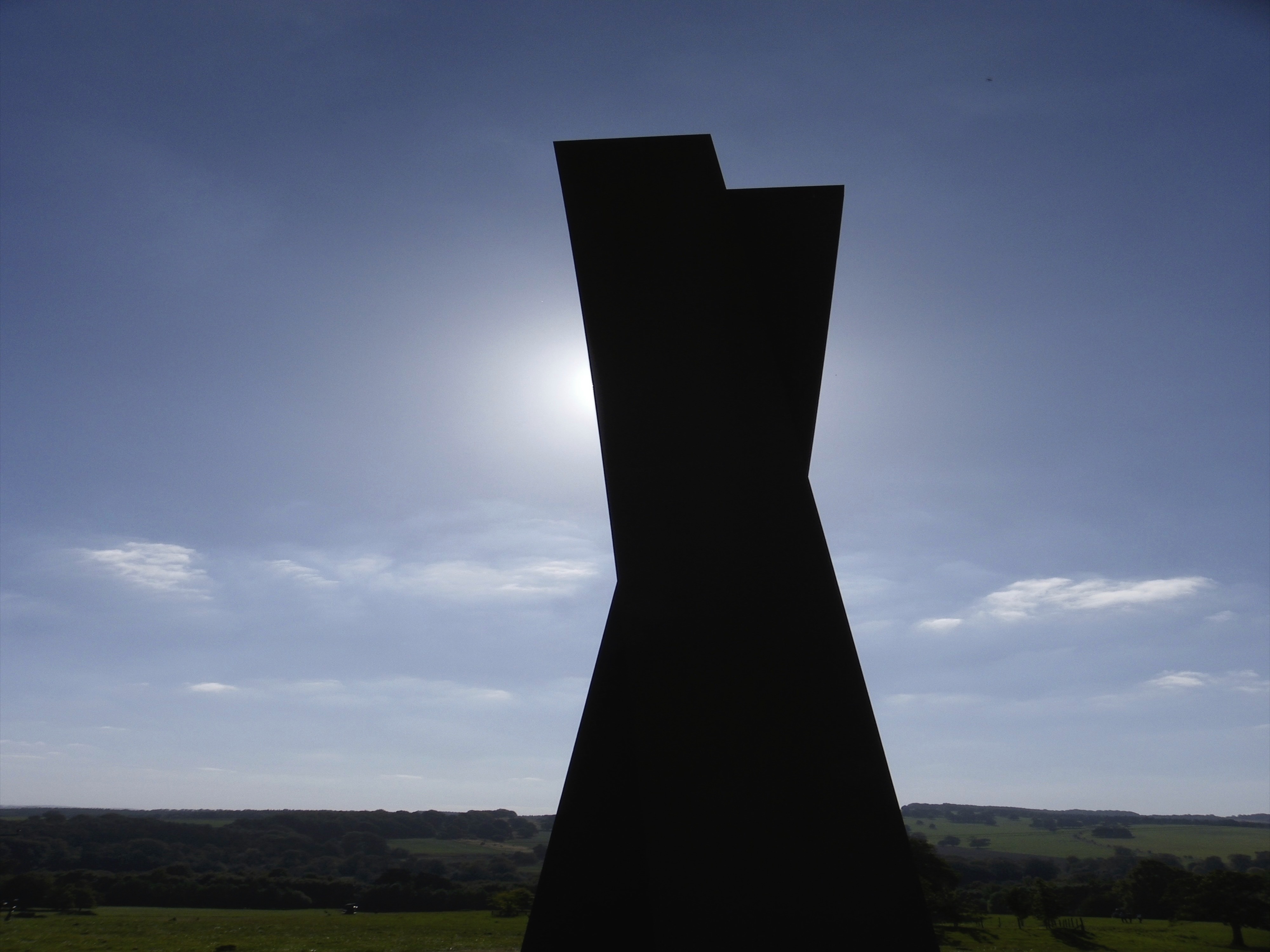 A silhouette of a a sculpture - abstract of two large blocks leaning against each other - with the sun high in the background.