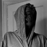 A hooded figure, bearded, hooded, stares at you.